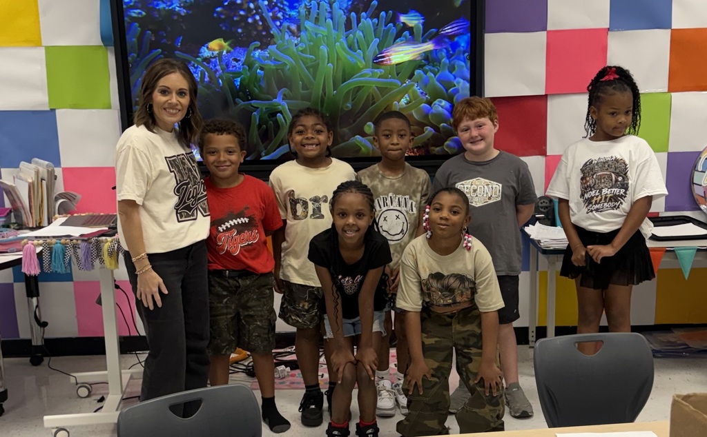 A group of children stands in a classroom, smiling for a photo. A woman in the middle wears a white shirt. A large aquarium is in the background.