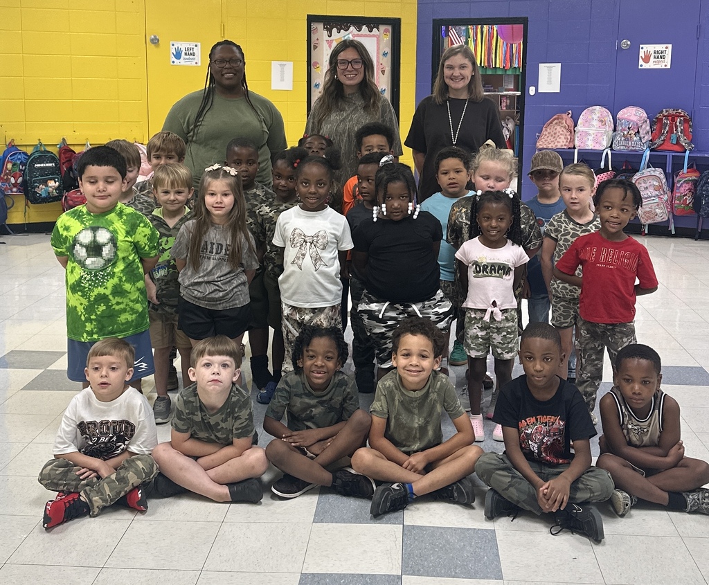 Group of children and two adults inside a classroom. Background features yellow and purple walls and backpacks.
