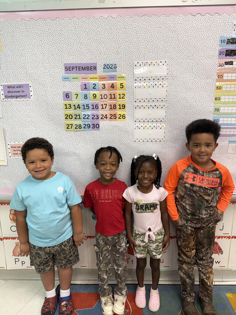 Four children stand in front of a classroom calendar for September 2023. The children wear casual attire.