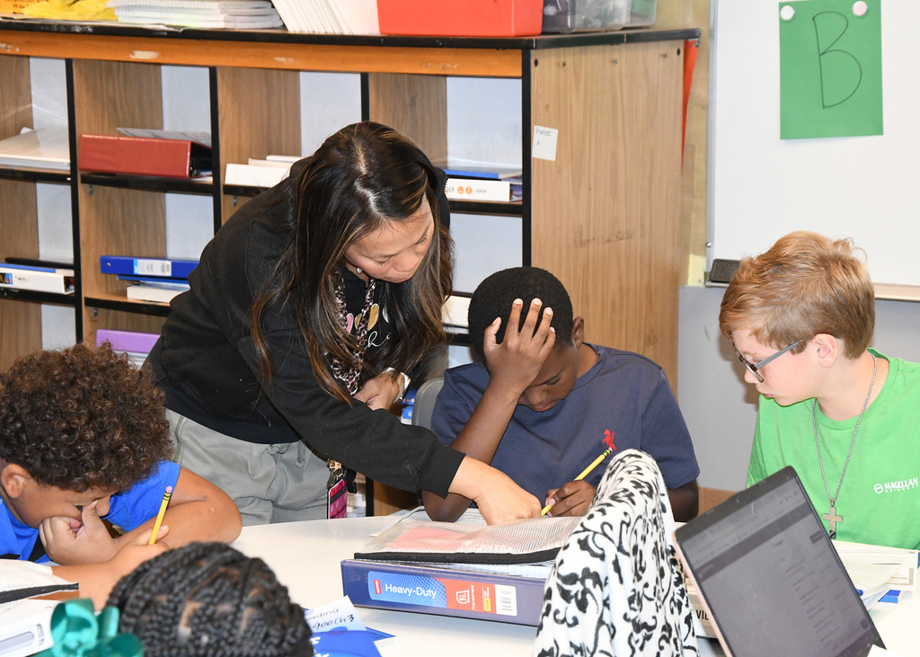 A woman helps two students with their homework. The students sit at a desk with a laptop and books.