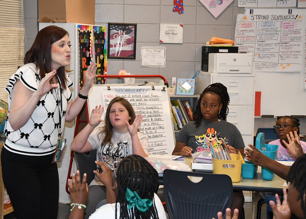 A teacher in a classroom gestures with hands raised. Several students in front, seated at a table, mirror her gestures.