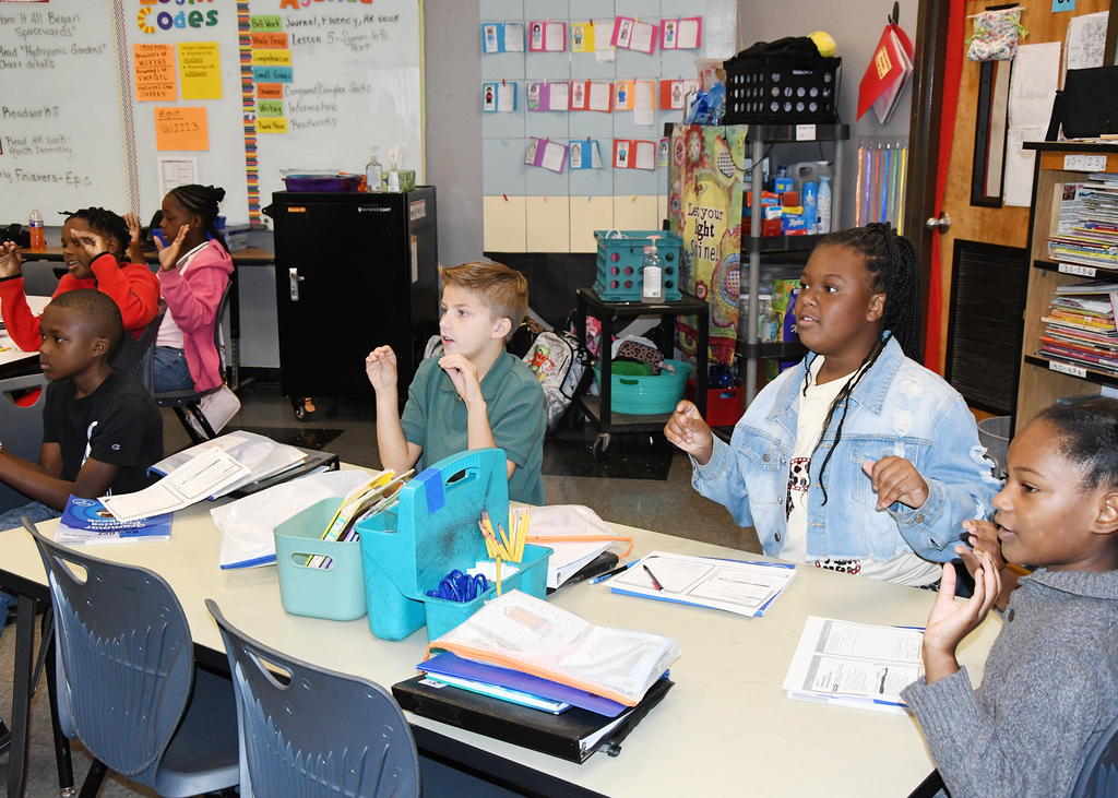 A classroom with students at tables. A girl with dark hair sits at the front, others around her.