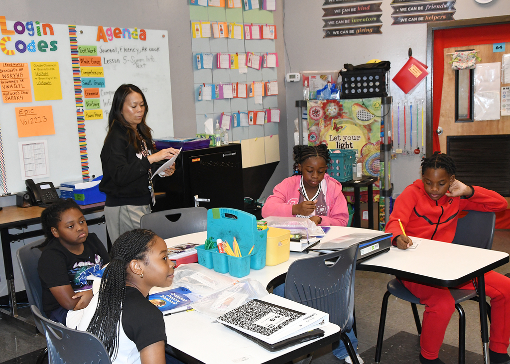 Teacher stands by classroom wall, facing students at tables. Four students work on assignments. Whiteboard with text and bulletin board with papers.