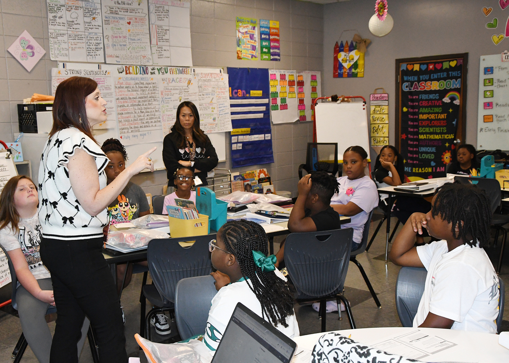 A teacher addresses students in a classroom. Desks, chairs, and educational materials are present. Brightly colored charts adorn the walls.