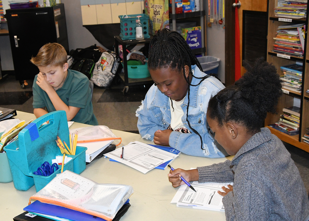 Three students at a table. One looks at a book. Another writes in a notebook.