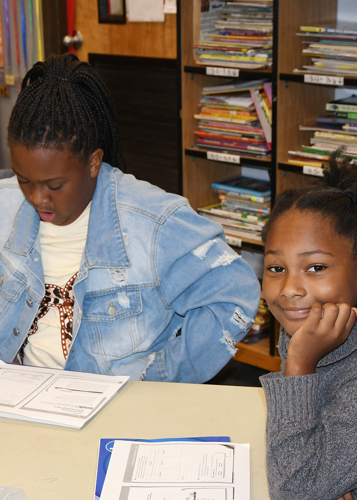 Two girls sit at a desk, one focused on a book and the other smiling, with bookshelves filled with books behind them.