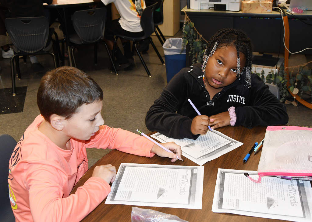 Two children, one in pink, one in black, sit at a table doing writing activities.