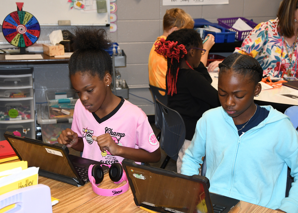 Two young girls sit at a table, each using a laptop. One wears a pink shirt and headphones, the other in blue.