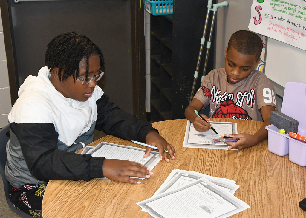 Two children work at a desk, one with glasses, one without, both holding pens and writing on papers.