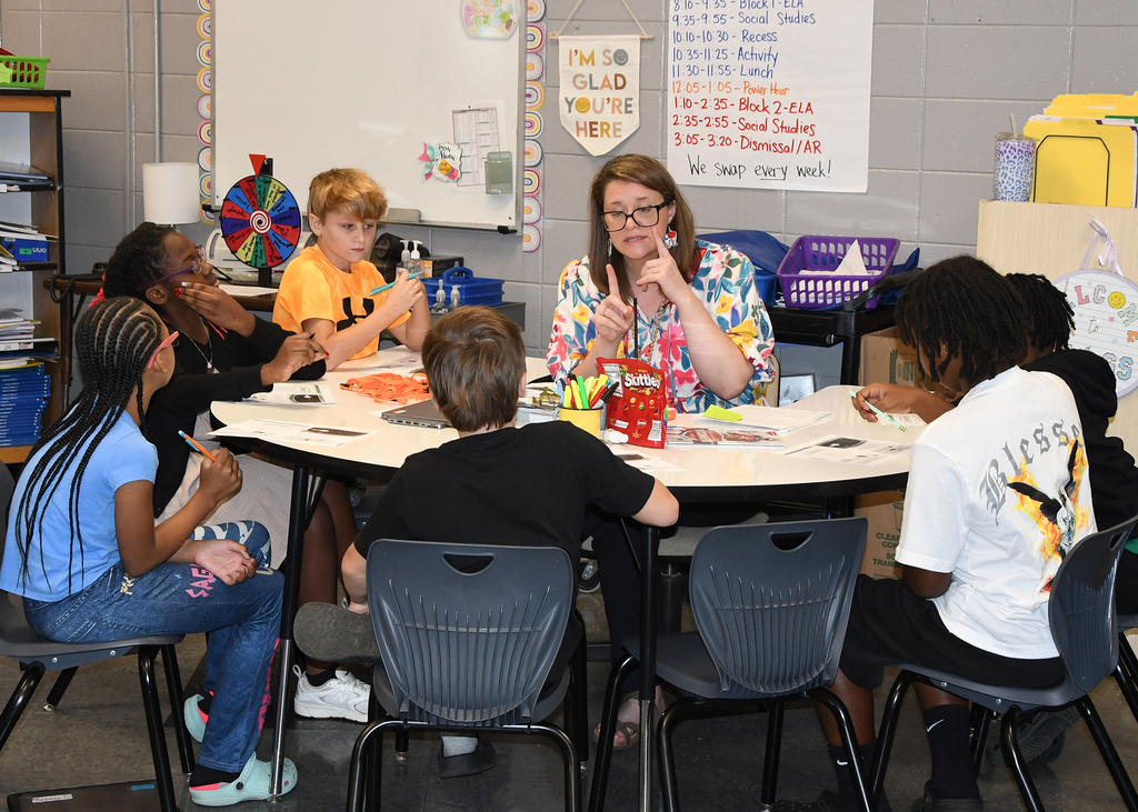 Classroom with teacher and students seated around a table, engaging in activities. Whiteboard and various classroom materials visible.