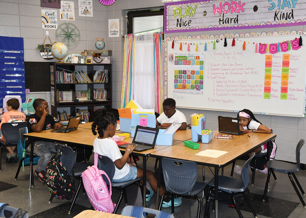 Students in a classroom with computers at tables. Bulletin board behind them displays "Play Nice, Work Hard, Stay Kind."