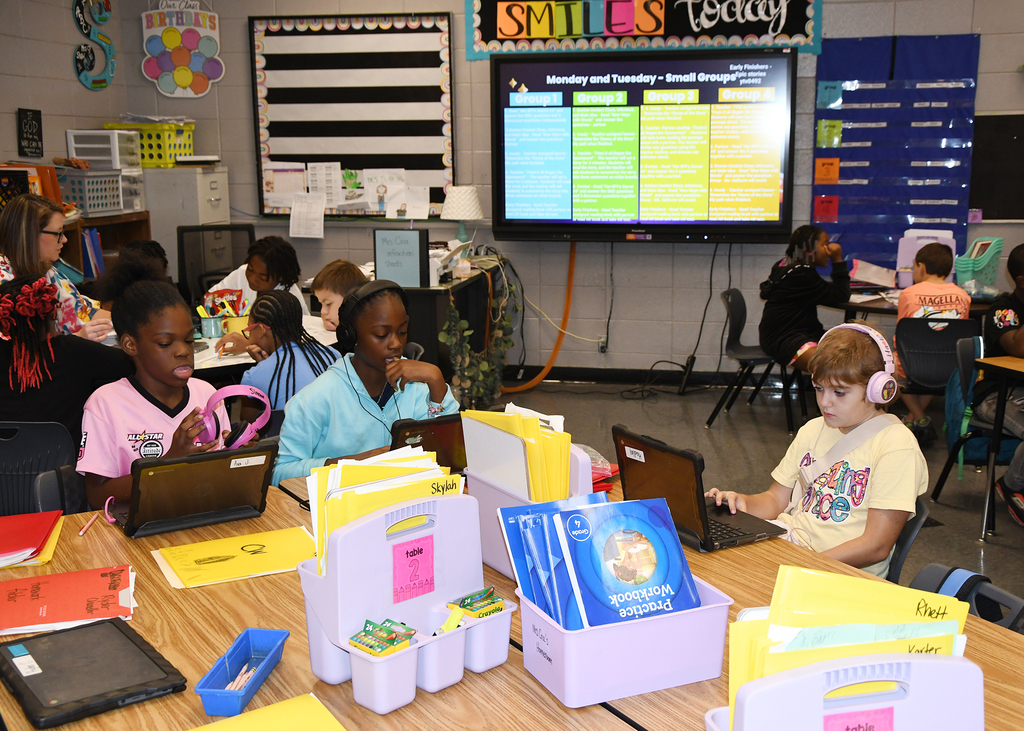 A classroom with children sitting at desks with laptops, folders, and books. A teacher monitors them in the foreground.
