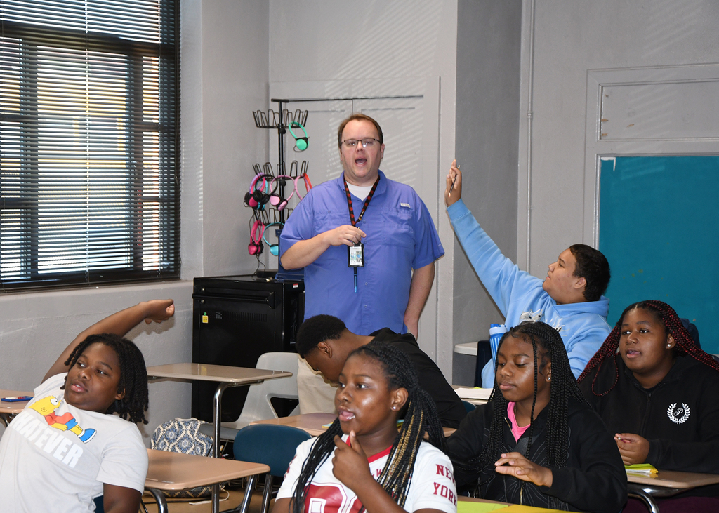 A teacher stands before students in a classroom. Students are seated, some raising their hands. Background includes a window, door, and a whiteboard.