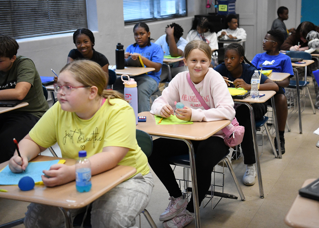 Classroom with desks, students wearing casual clothes, engaged in activities. A girl in yellow smiles.
