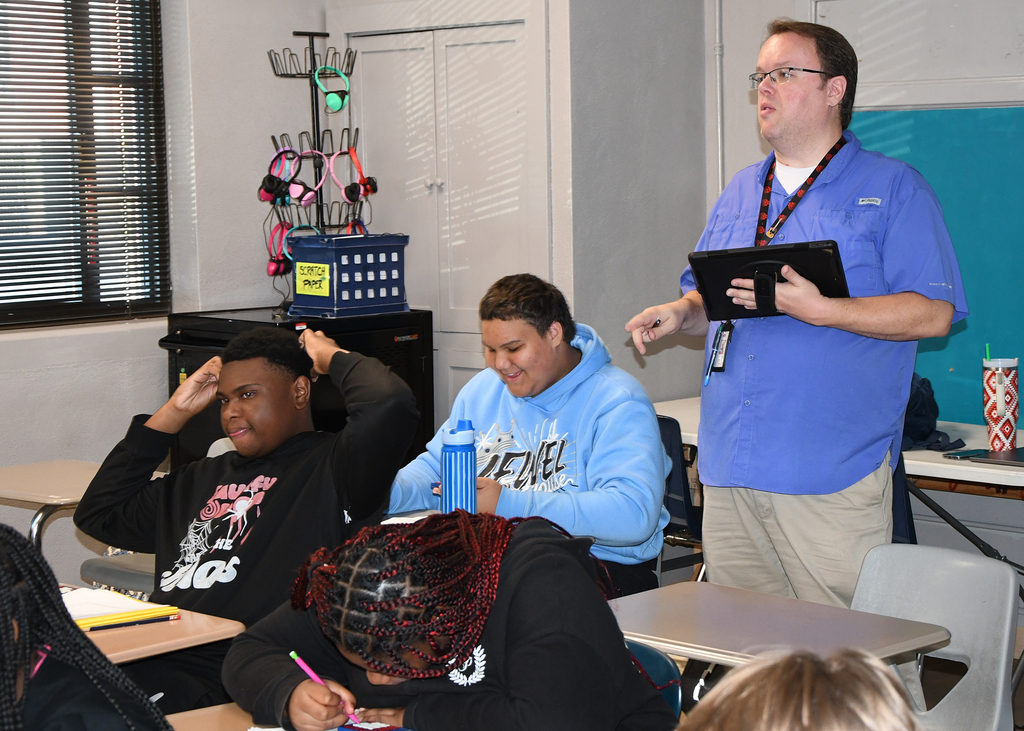 Teacher standing in classroom with students. One student is seated with head down.