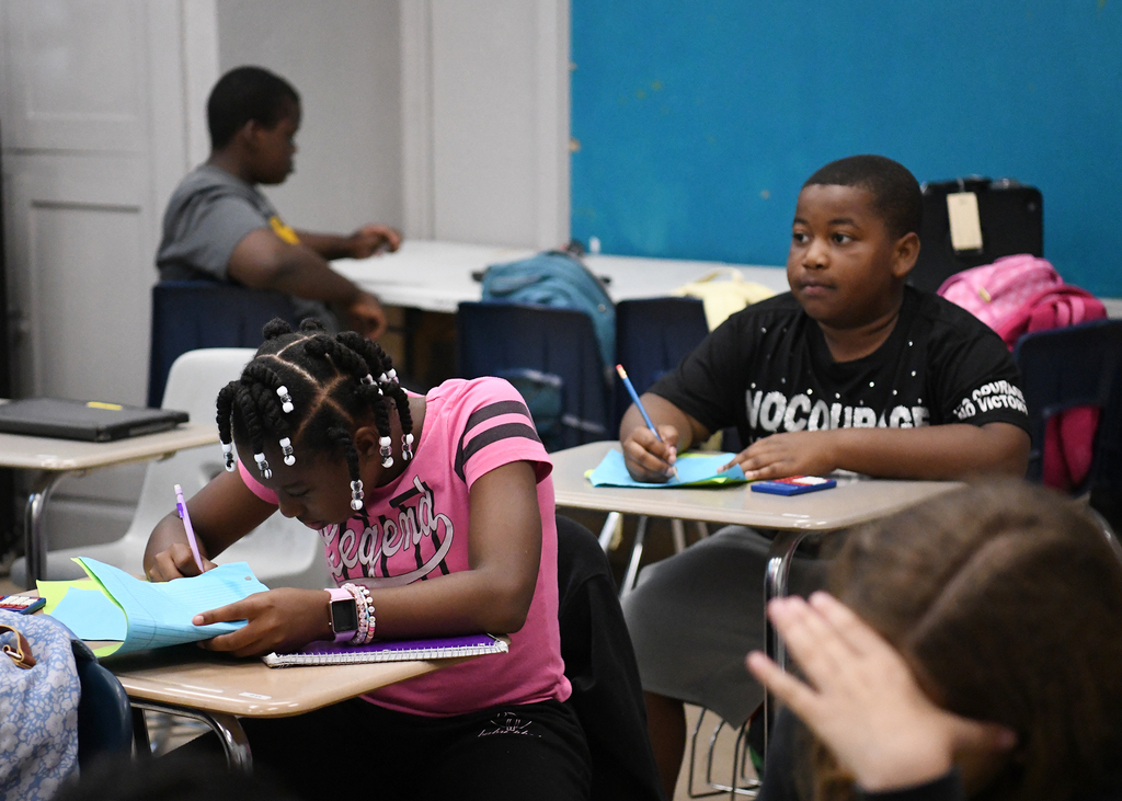 Students seated at desks in a classroom. One girl in pink focuses on her notebook. A boy in black writes beside her.