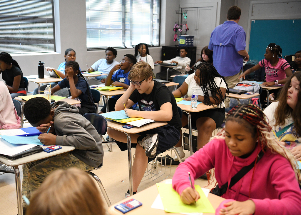 A classroom filled with students sitting at desks, some using books and papers, and a teacher standing.