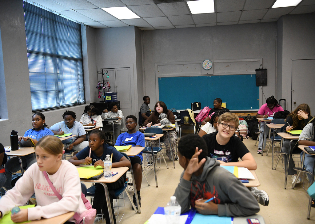 A classroom with students seated at desks. Some wear backpacks; others engage in conversation.