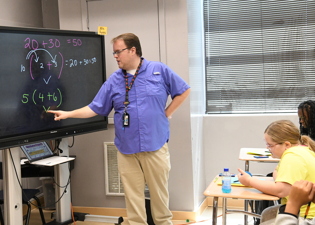 A man points to a whiteboard with math equations in a classroom. A student sits at a desk, listening.