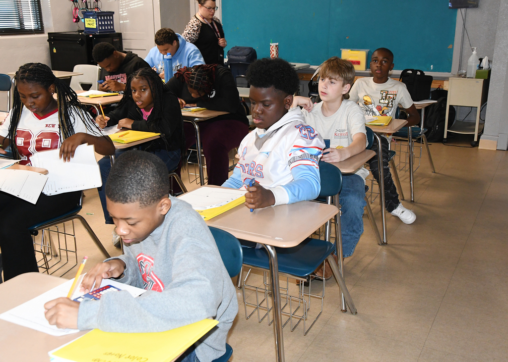 A classroom with students in desks. A boy in a gray shirt writes on a paper. A girl in red holds papers.
