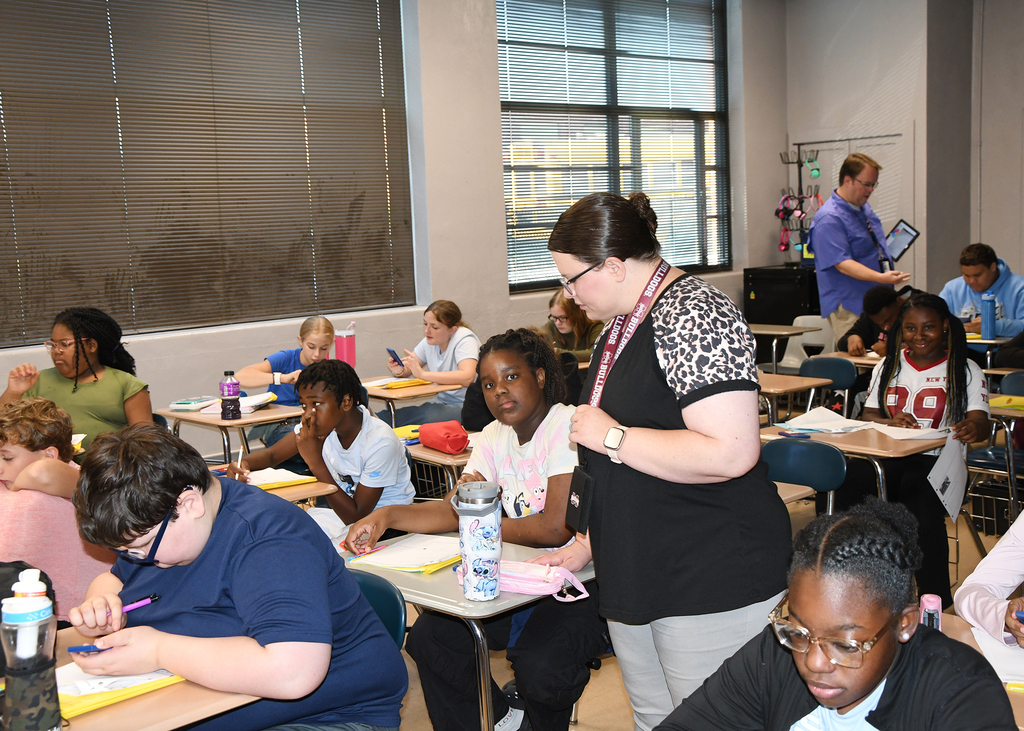 A classroom with students sitting at desks; a woman stands at the front, likely teaching, with large windows in the background.