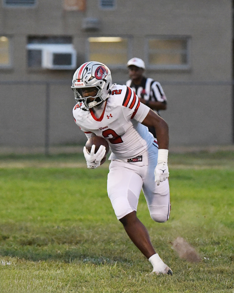 A player runs with a football on a grassy field. A referee watches in the background.