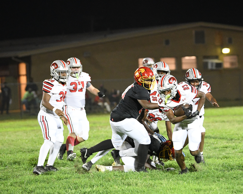 Football players in jerseys are running on the field. One player tackles another. An audience watches from the sidelines.