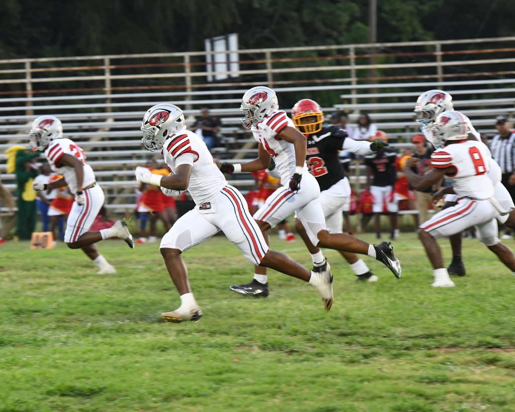 Five football players in white and black uniforms run across a field. Two players wear red helmets.