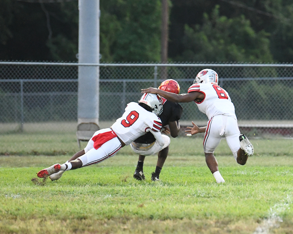 Players on a field with two wearing white and red uniforms tackle a player in a black uniform.