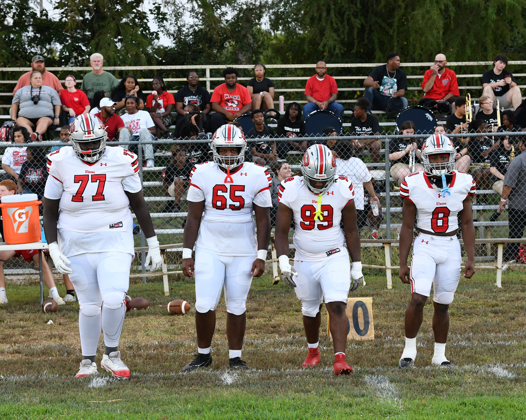 Four football players in white uniforms walk together on a field. Behind them, spectators sit on bleachers.