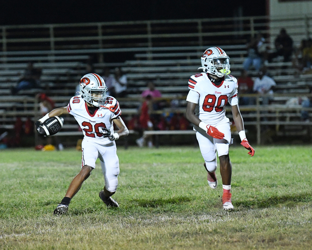 Two football players run on a field. One holds a football, while the other chases him. Spectators watch from bleachers.