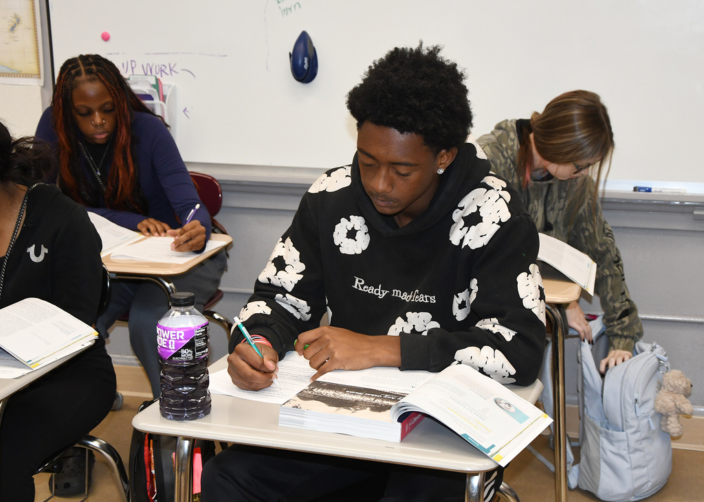 Four people sitting at desks in a classroom. One is writing with a pen and the others are engaged in various activities.