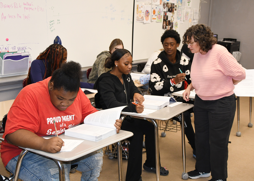 In a classroom, students work on papers at desks. A teacher assists one student, holding a pen.