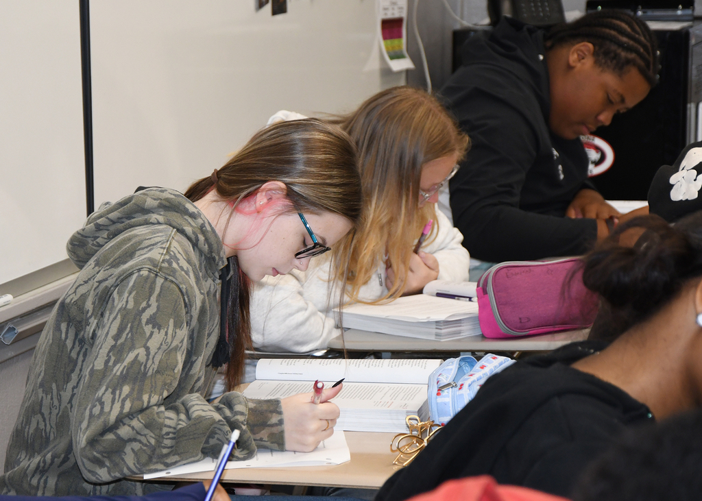 Several students write in a classroom, two with heads bent down, one with eyes closed, and another focused on writing.