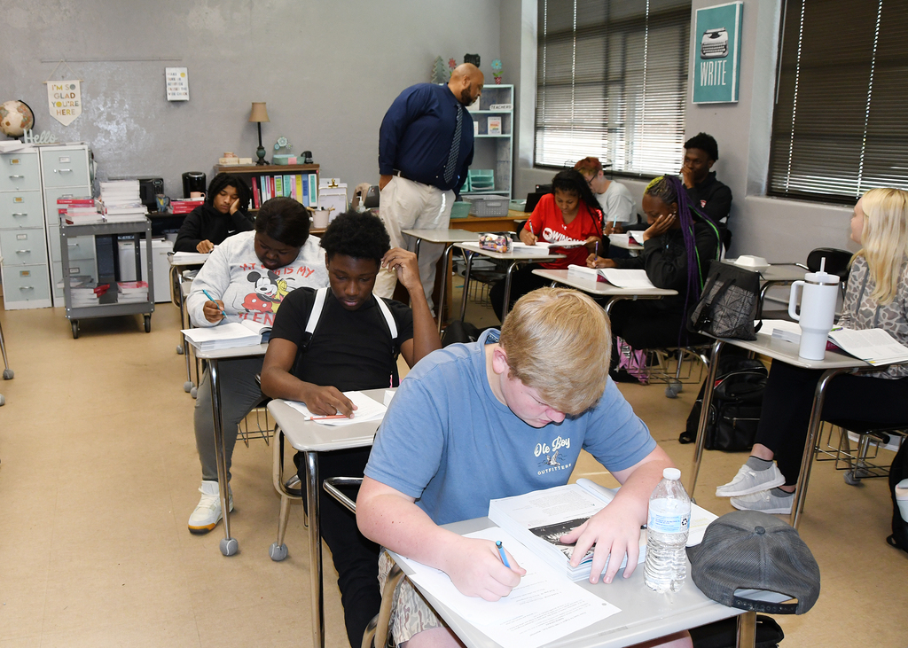 Students in a classroom with desks, one student leans over a desk to write while others sit.