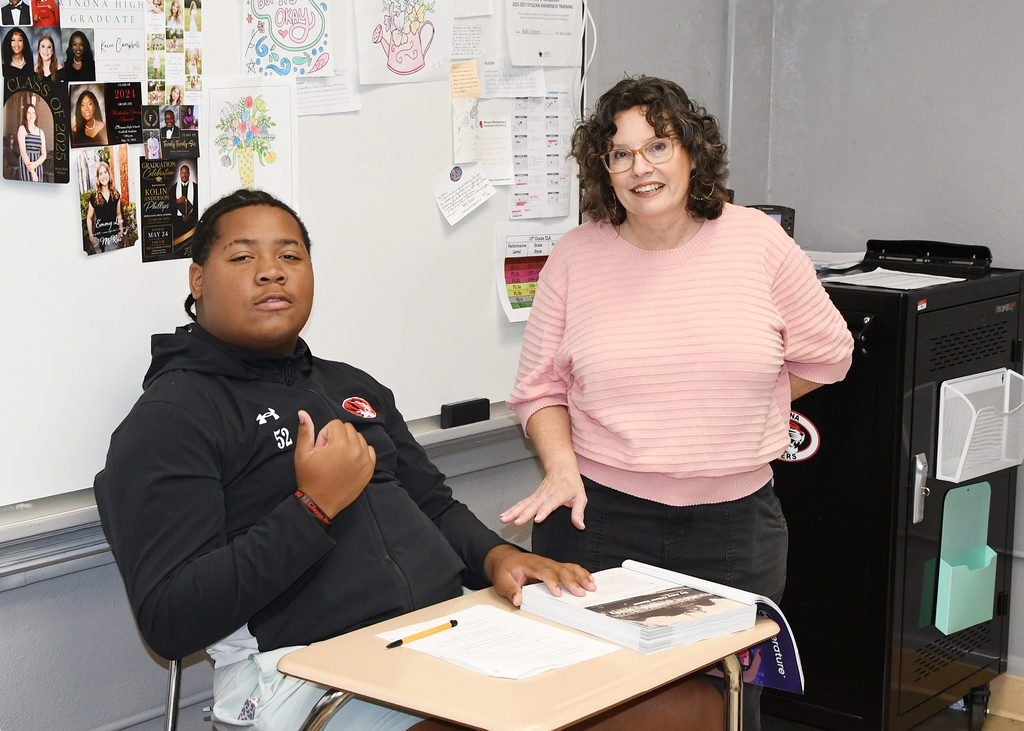 A young man sits at a desk while a woman stands behind him, both facing the camera. The background features a whiteboard with various papers and pictures attached.