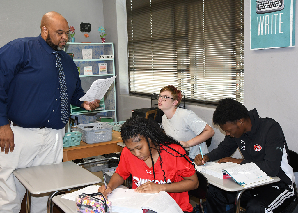 A teacher stands before a group of students seated at desks. One student writes while others look on.