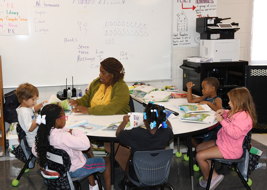 A classroom with a teacher and six students seated at a round table. The teacher is seated at the head of the table, while the students sit in chairs.