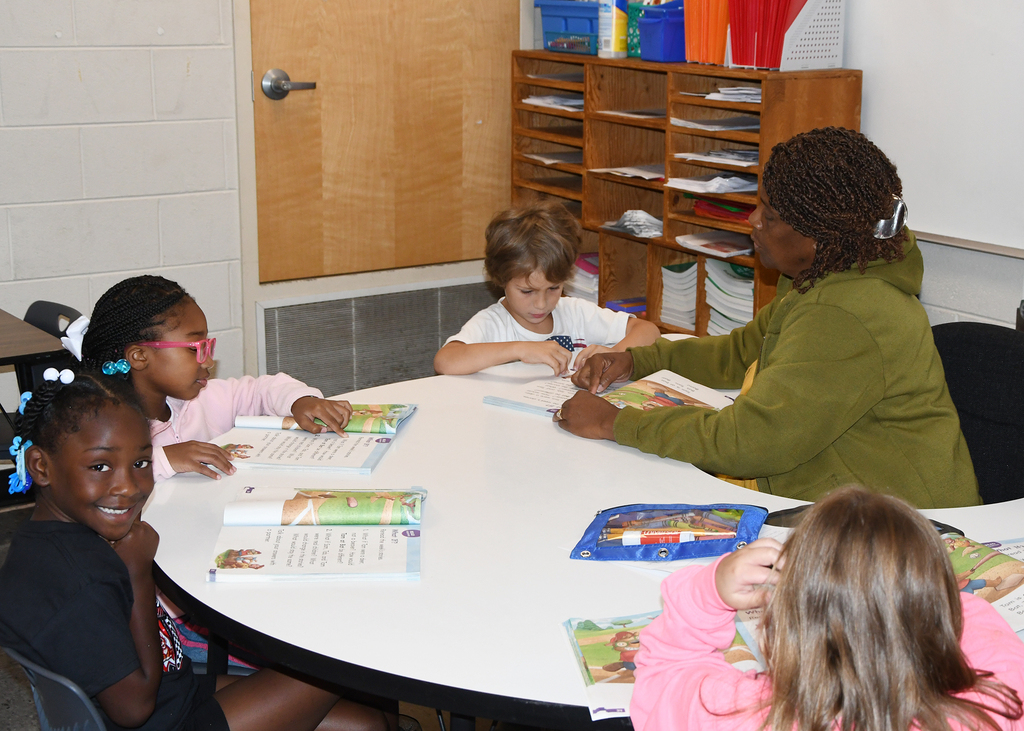 A classroom with children seated at a round table, reading books. A teacher is seated next to them, assisting with reading.