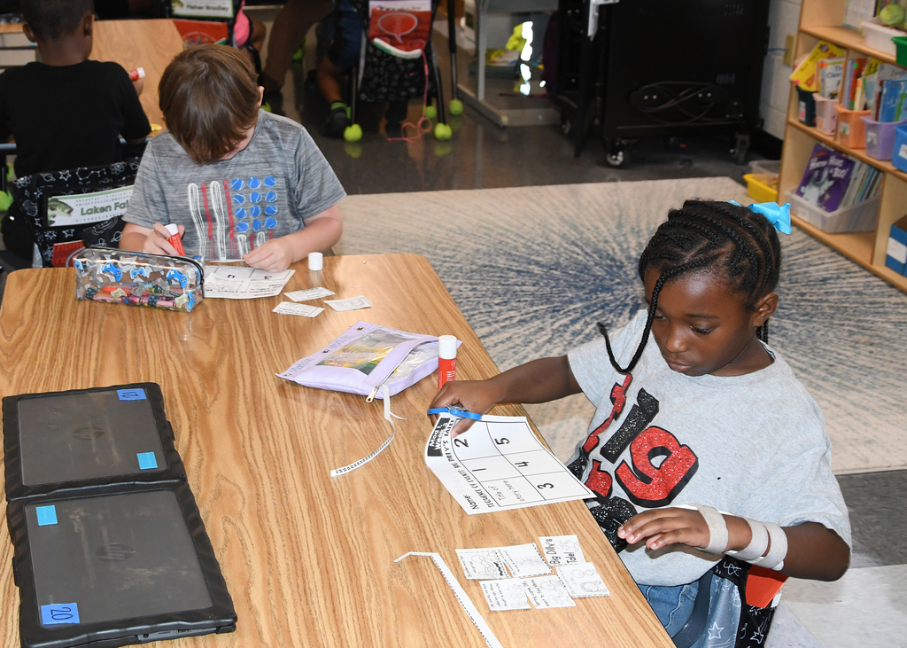 Two children seated at a table work on tasks. One has papers and a pen. The other has a tablet.