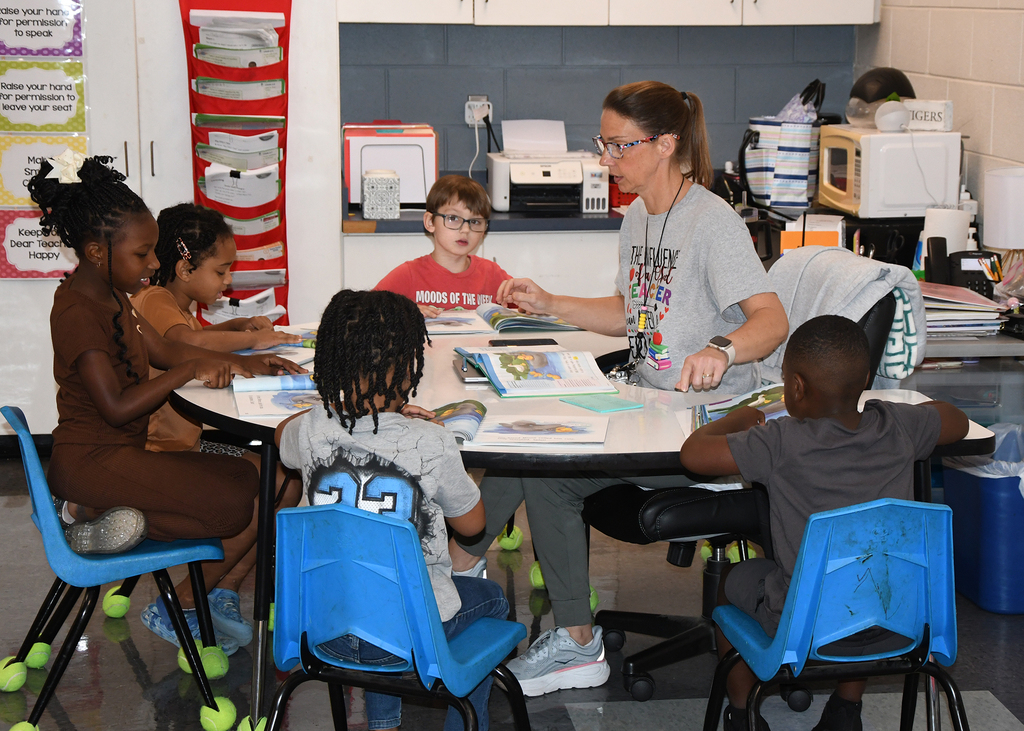 An instructor guides a group of children seated around a round table, surrounded by colorful classroom decorations.