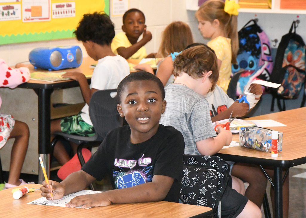A classroom with children seated at desks, one child smiles while writing, and several others work at their tables.