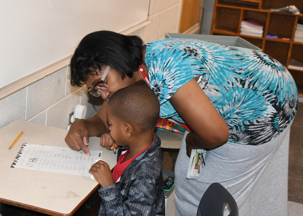 Woman wearing glasses and a patterned shirt helps a young boy with his schoolwork at a desk.