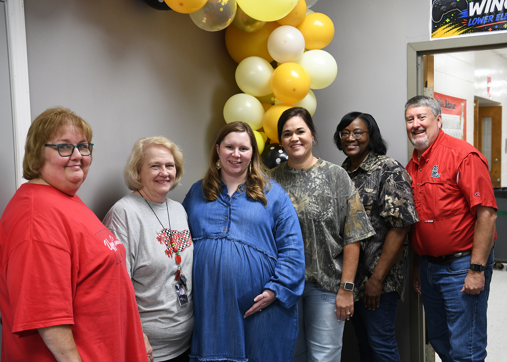 Six adults stand by a wall with a large balloon display. One woman wears a baby bump.