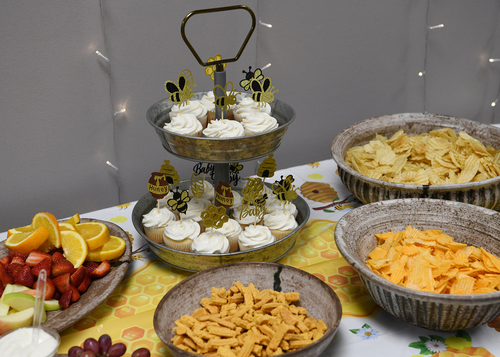 A table holds a tiered tray with cupcakes, bowls of chips, fruit slices, and a bowl of cheese.