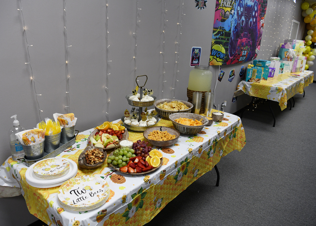A table with a yellow tablecloth displays various foods, including fruits, snacks, and desserts, with balloons in the background.