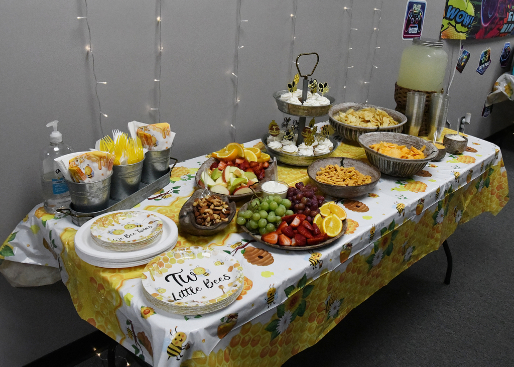 A table with a yellow floral tablecloth has various foods on it, including fruits, cheese, nuts, and a cake.