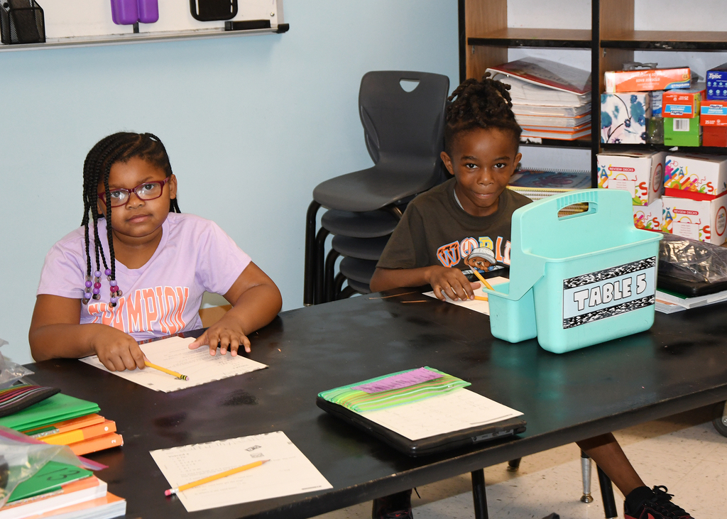 Two young girls seated at a desk, each working on papers. One wears glasses, and the other wears a gray shirt.
