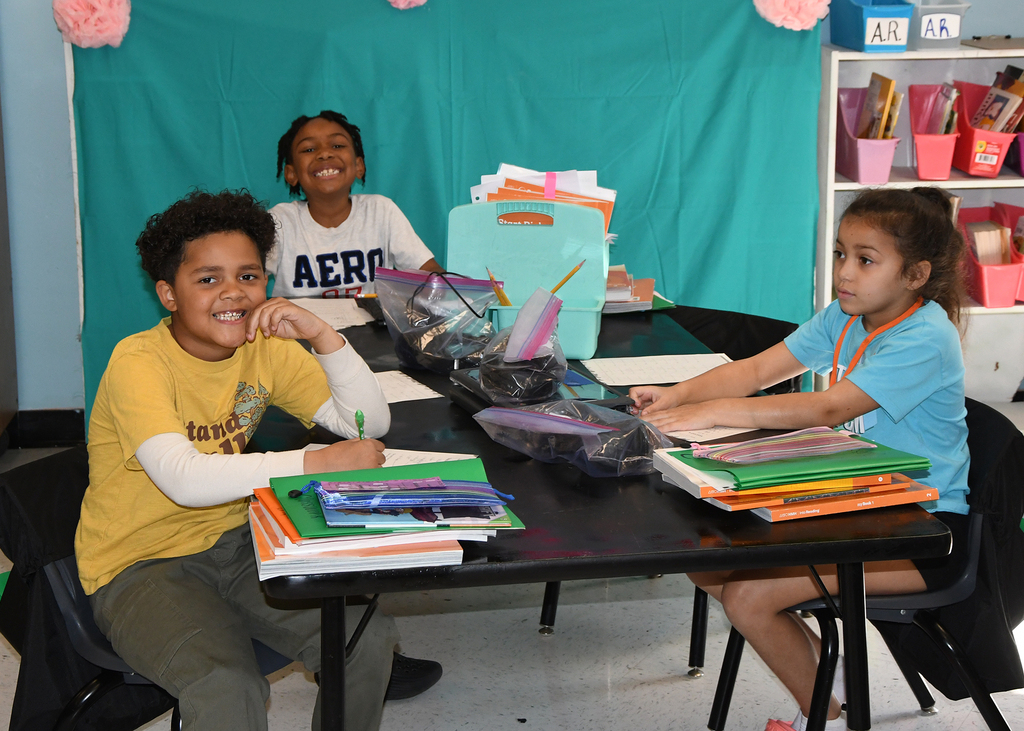 Three children sitting at a table, smiling and working with school supplies, with a teal backdrop.