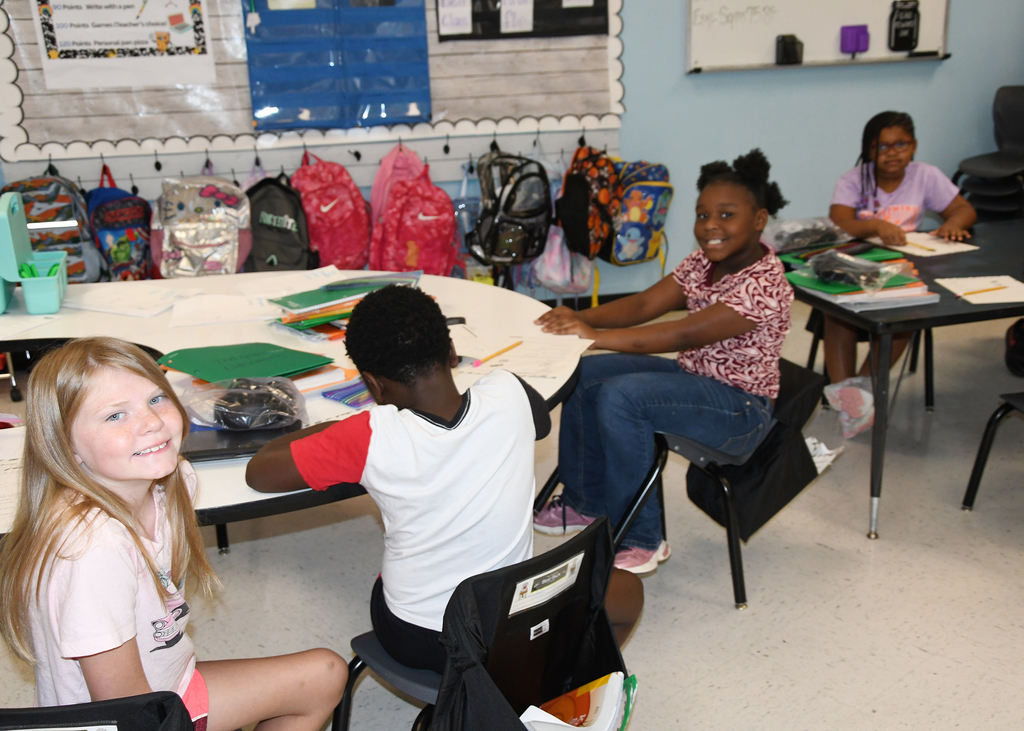 Four children seated at tables in a classroom, with papers, pencils, and bags on the desks.